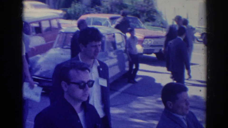 1967 Los Angeles frame showing attendees with sunglasses and name tags walking near parked classic cars in daylight.