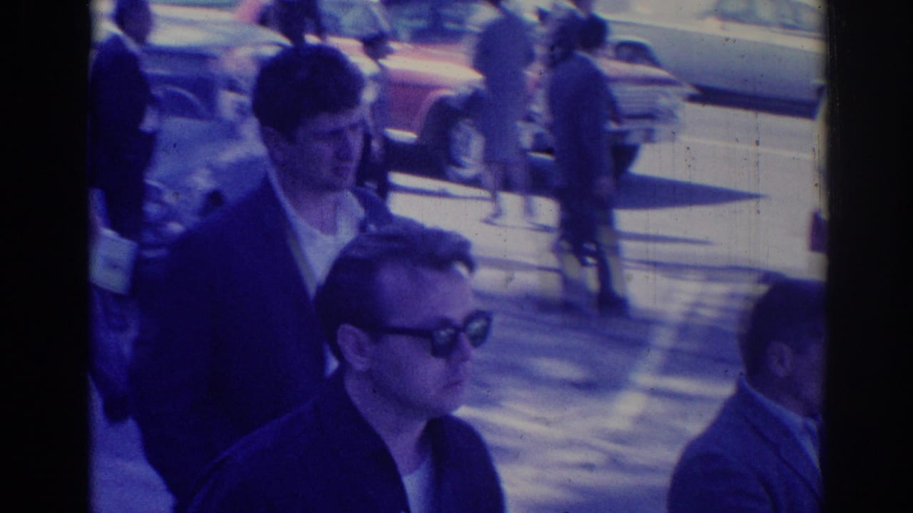 Adults walk on a sunny city sidewalk near parked cars, with one man wearing dark sunglasses.