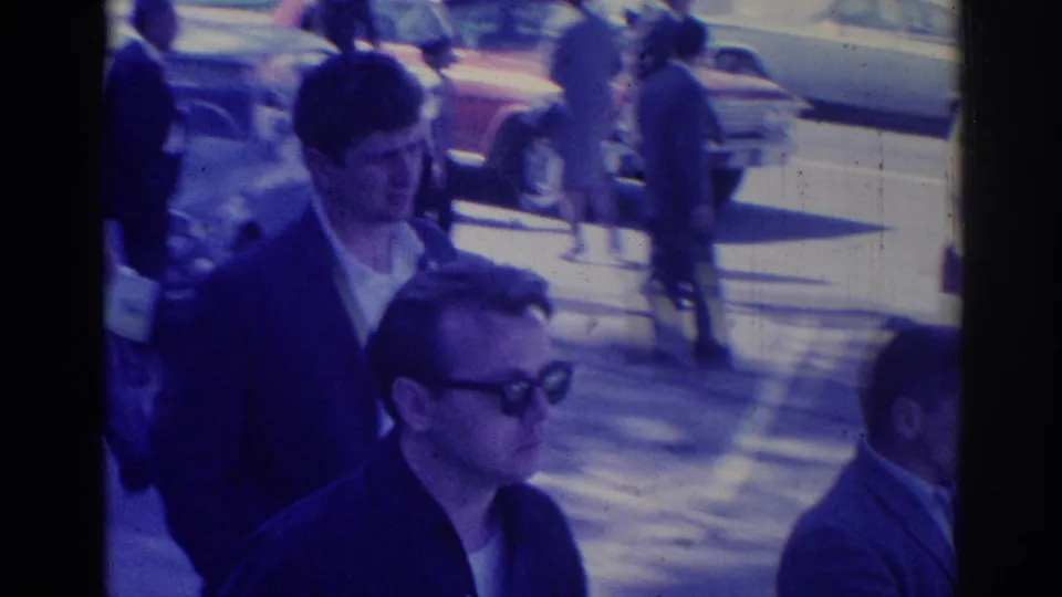 1967 Los Angeles home-movie frame showing adults walking through a busy sidewalk scene near classic parked cars in bright sunlight.
