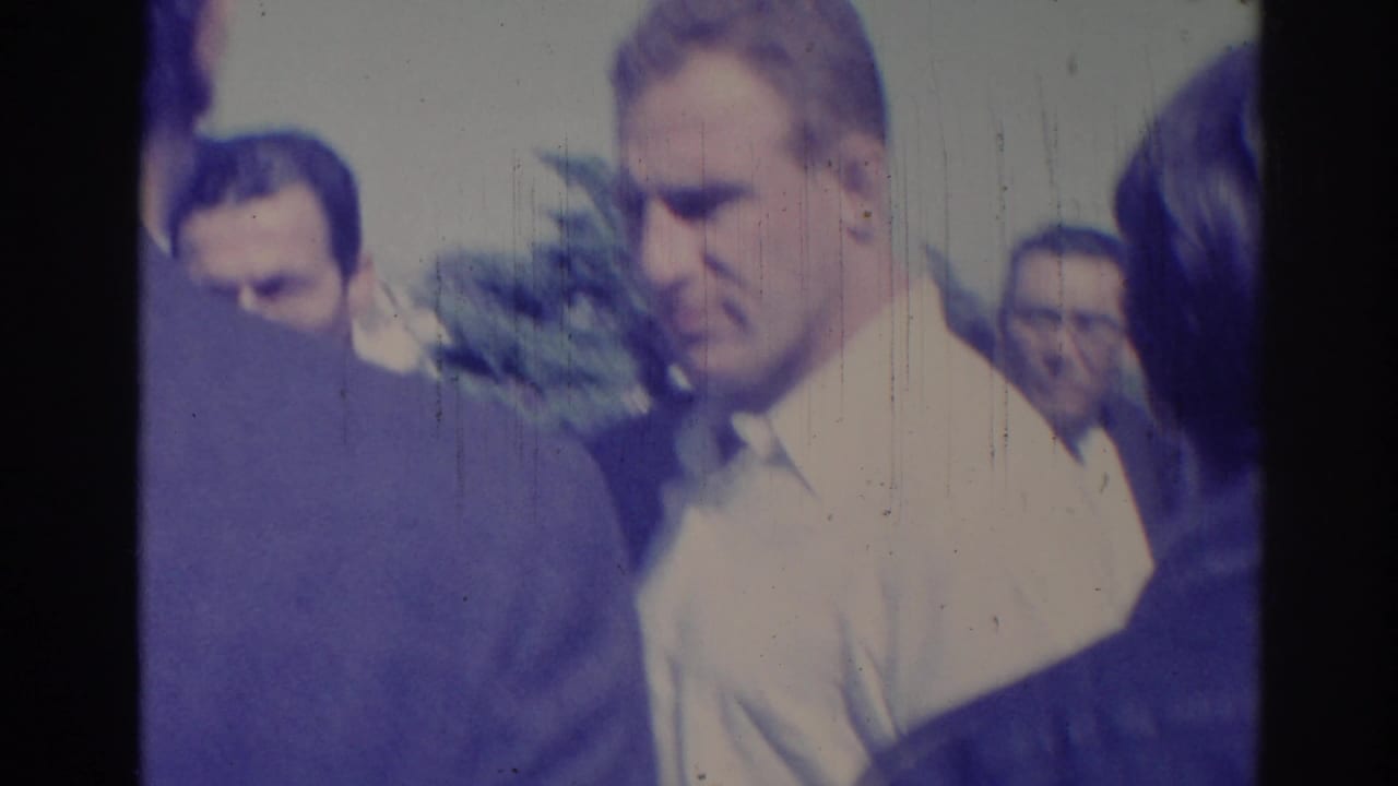 Close handheld 1967 Los Angeles street view of adult men gathered outdoors in bright light, with faces partly soft from the 8mm scan.