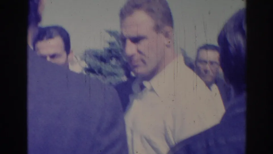 Close handheld 1967 Los Angeles street view of adult men gathered outdoors in bright light, with faces partly soft from the 8mm scan.
