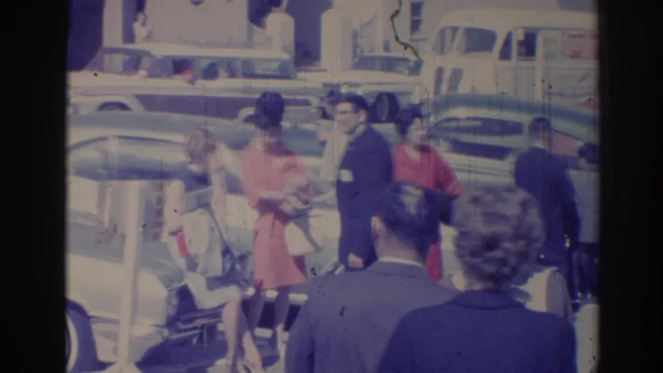 Wide 1967 Los Angeles street frame with adults crossing a busy sidewalk near mid-century cars, including a woman in a red dress.