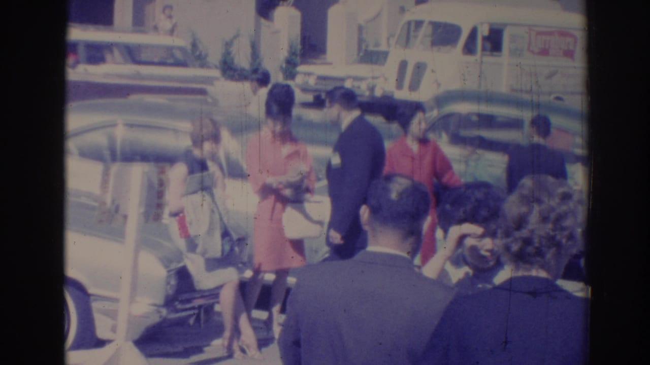 People gathered near parked cars and a white delivery van on a sunny street.