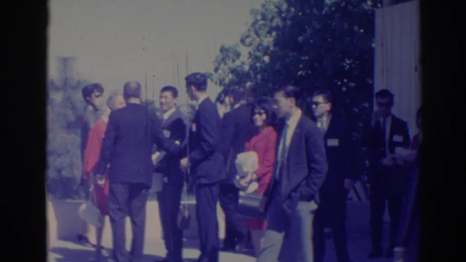 1967 Los Angeles home-movie frame of adults in suits and dresses gathered on a sunny sidewalk.