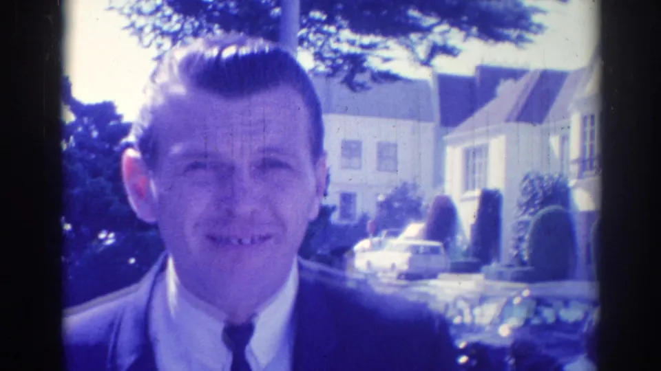 Clearer 1967 Los Angeles frame of a smiling suited man with suburban houses and parked cars behind him.