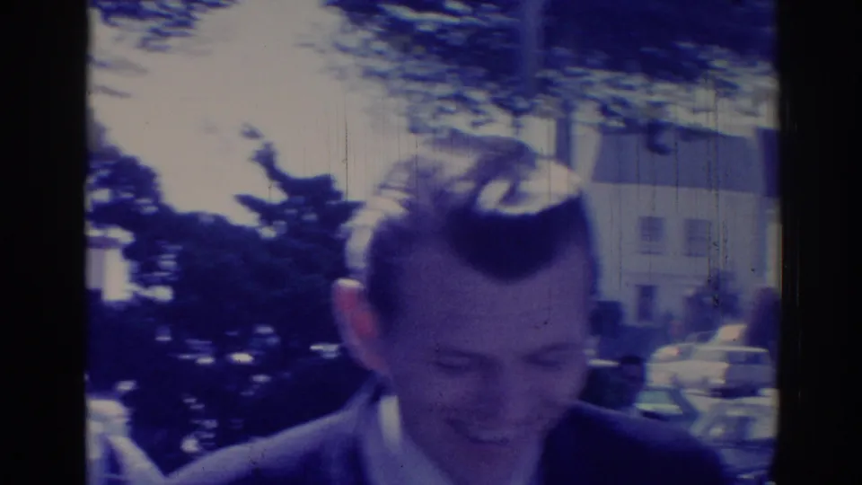 Handheld 1967 Los Angeles frame of the suited man looking down near trees, houses, and parked cars.