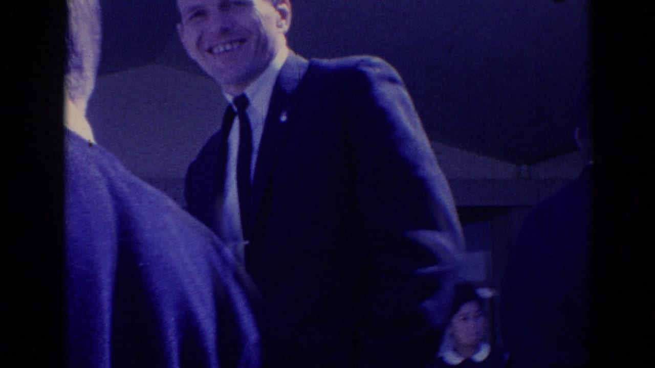 Smiling man in a dark suit standing close to another person outdoors.