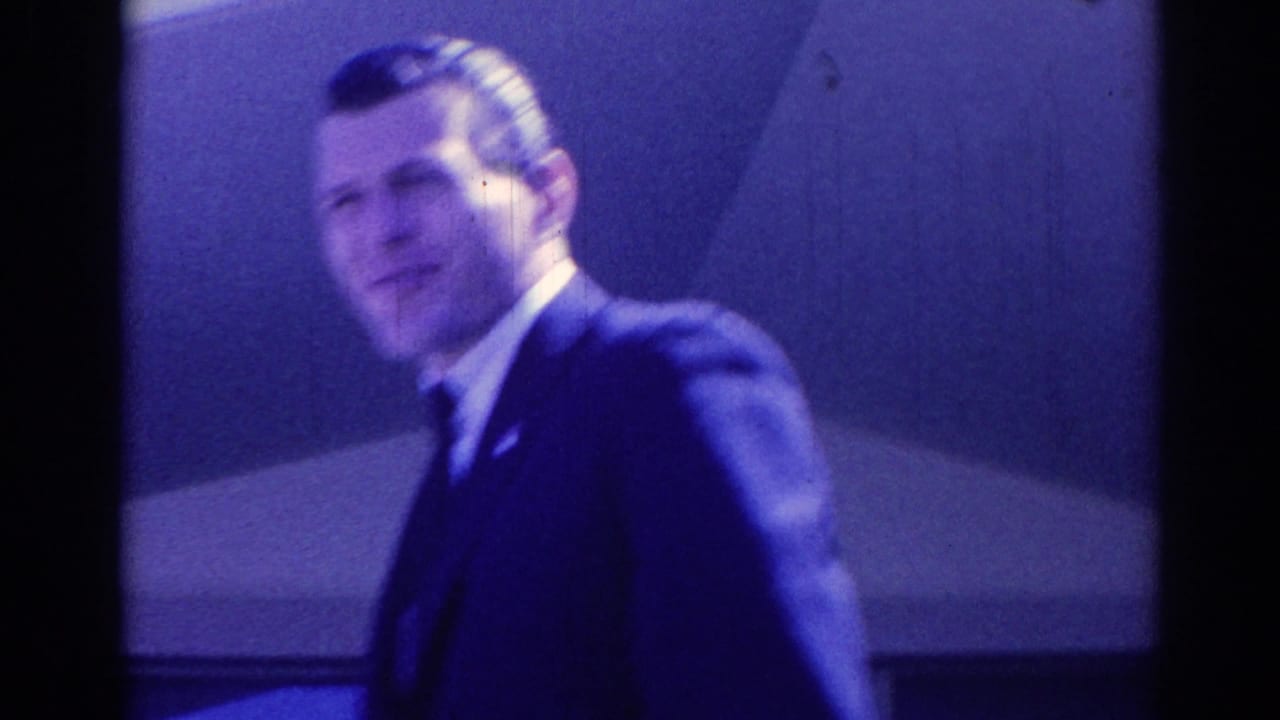 Man in a dark suit smiling toward the camera near a building wall.