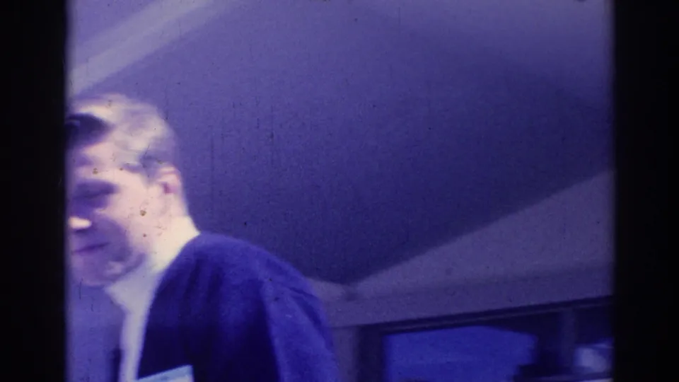 Soft 1967 Los Angeles home-movie frame of a suited man passing close to the camera under a building overhang.