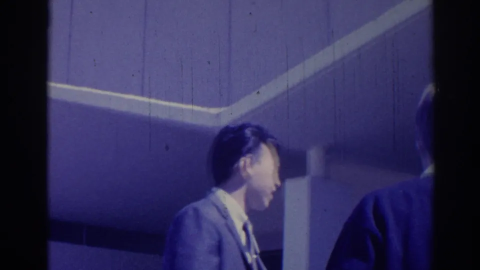 1967 Los Angeles frame with two suited men near a plain interior-looking wall, one in profile and one partly visible.