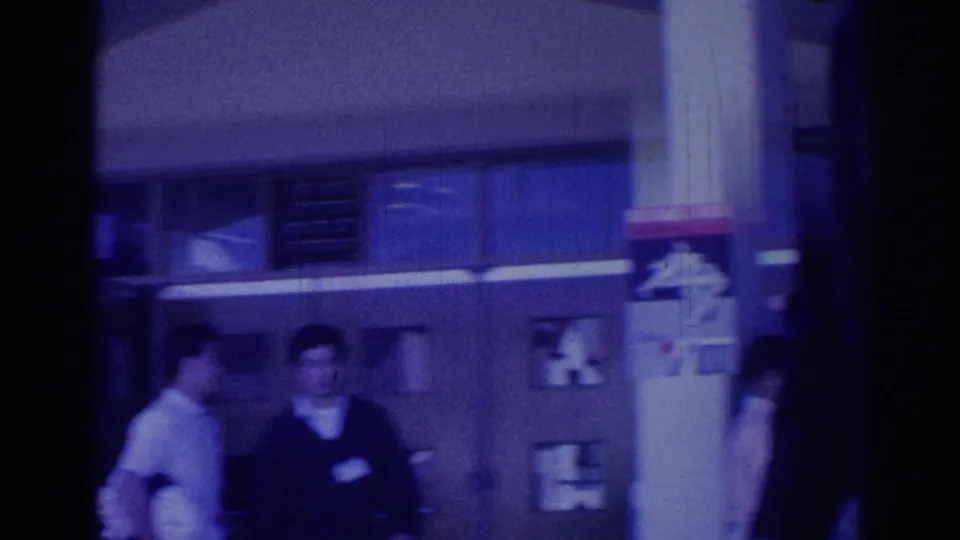 1967 Los Angeles frame with adults gathered near glass windows and a partially visible sign or emblem in a formal-event setting.