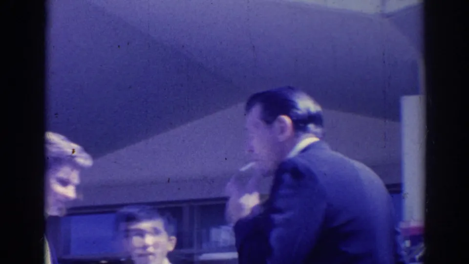 1967 Los Angeles frame showing adults grouped near a building wall, with a suited man in profile and faces partly visible.