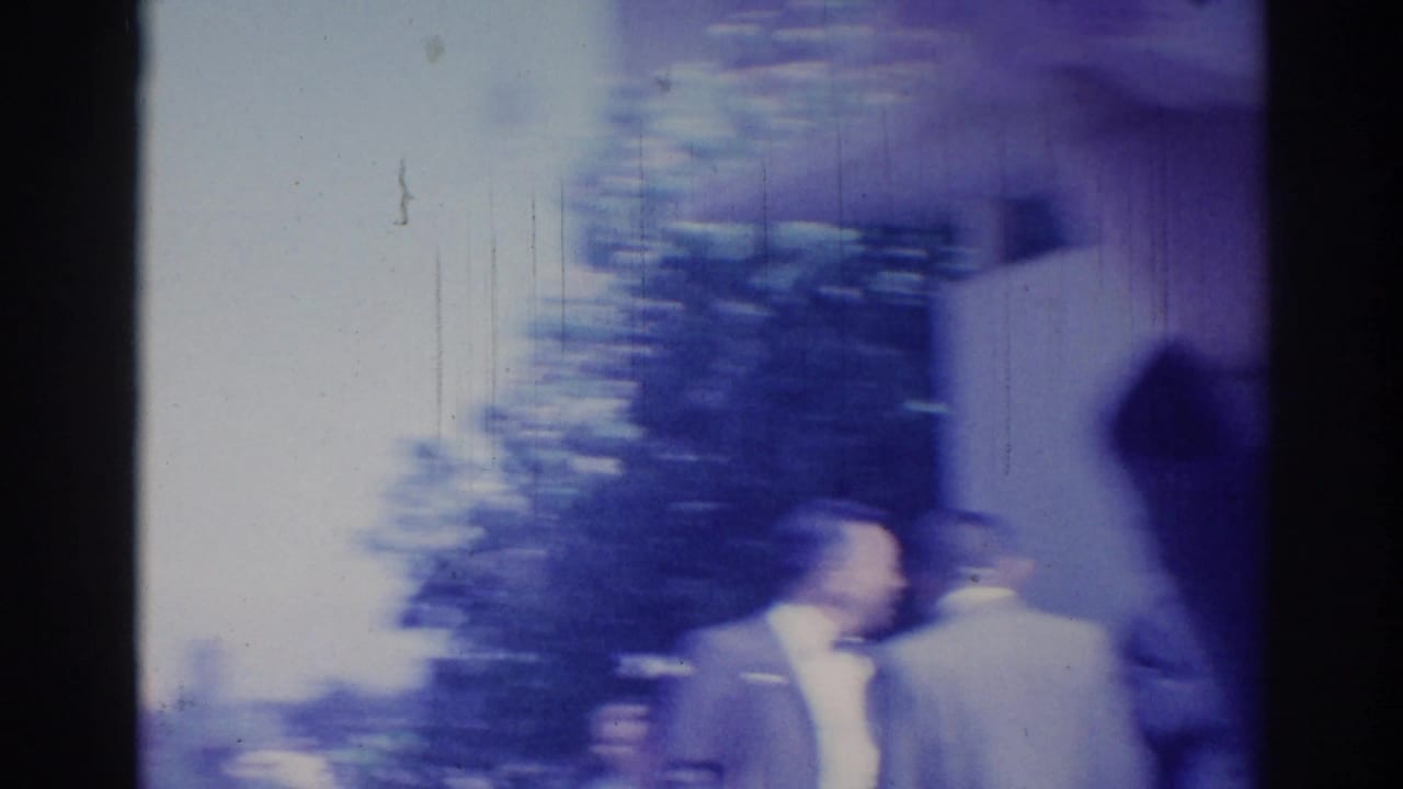 Handheld 8mm frame from the Los Angeles gathering, with blurred figures near a building entrance and trees.