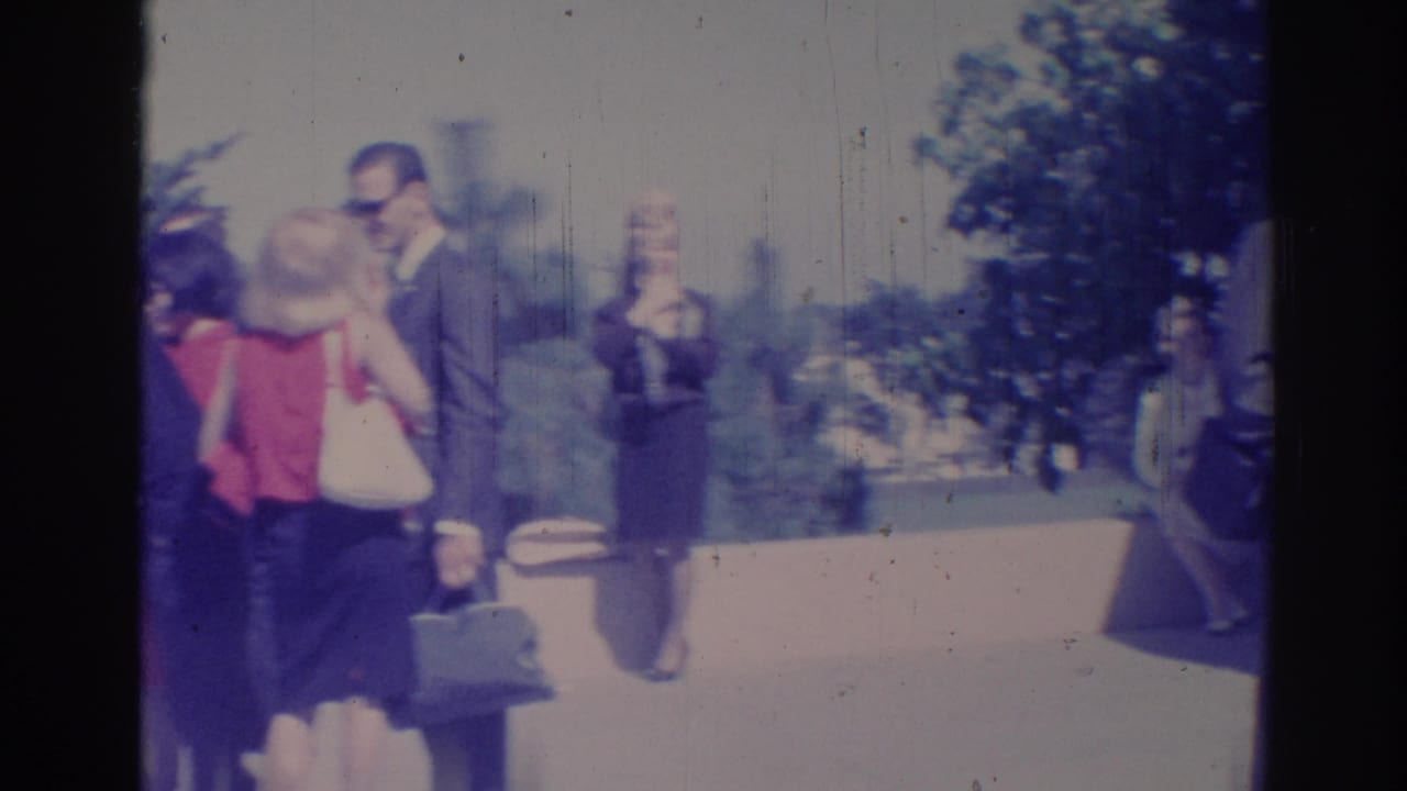 1967 Los Angeles home-movie view of a woman with a handbag near men in suits on a sunny outdoor walkway.