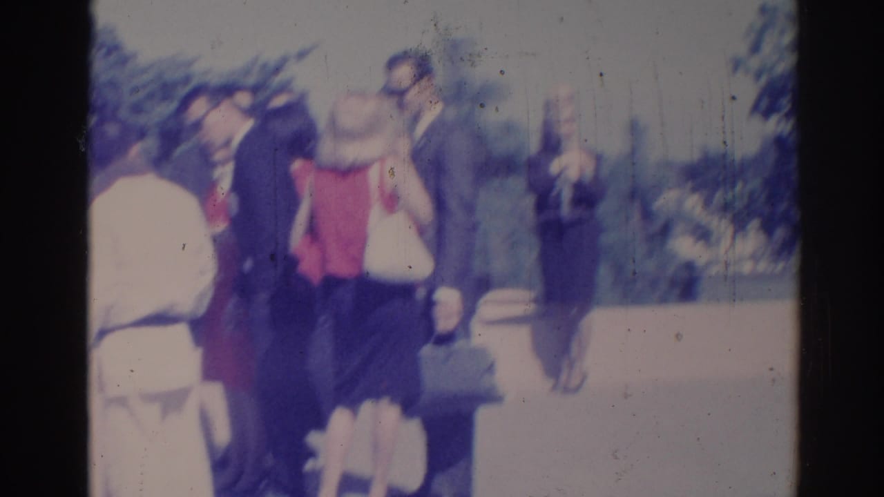 Soft 8mm frame of adults conversing outdoors in Los Angeles, with suited men and a woman in red visible.