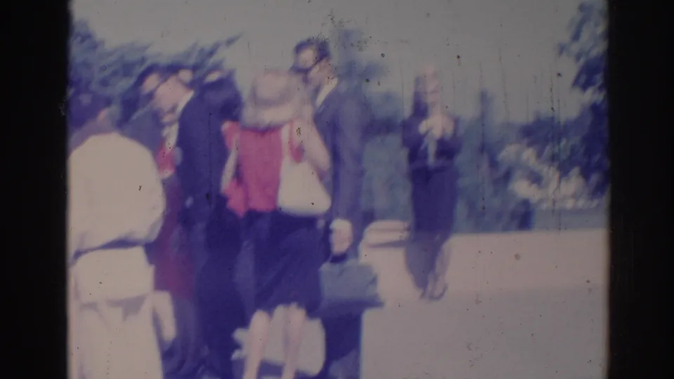 Soft 8mm frame of adults conversing outdoors in Los Angeles, with suited men and a woman in red visible.