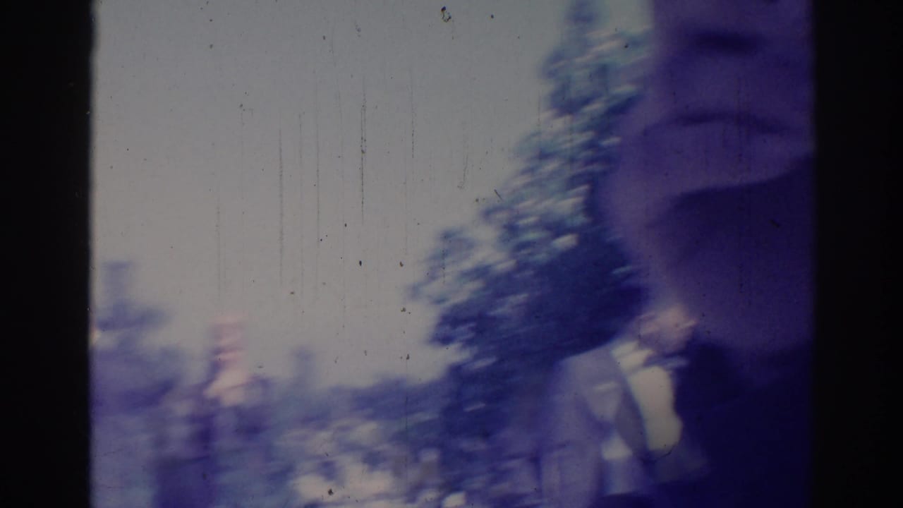 Blurred outdoor 8mm frame with an adult figure and trees or street shapes near the edge.