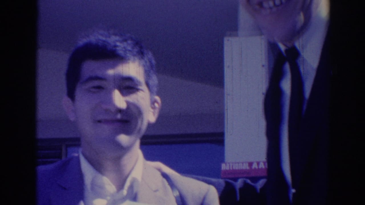 Young man in a light suit smiles at the camera in 1967 Los Angeles, with another suited adult partly visible behind him.