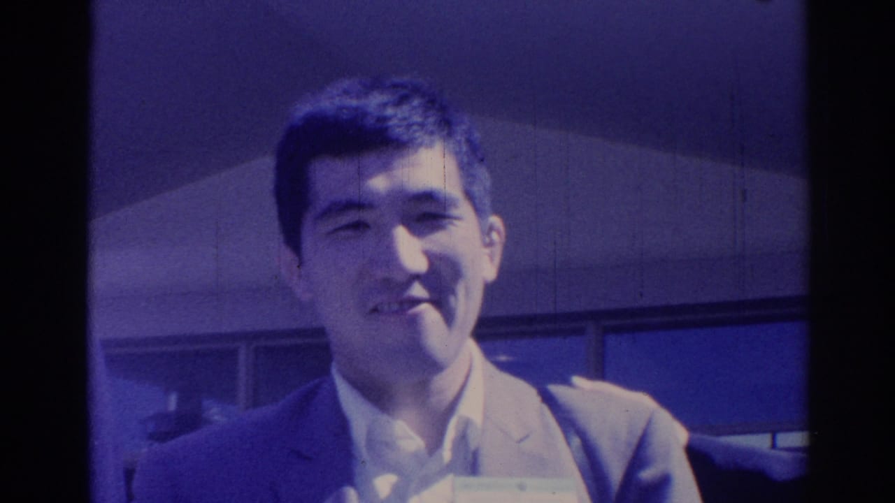A young man in a light suit smiles outdoors in this 1967 Los Angeles 8mm home-movie frame, with a building facade behind him.