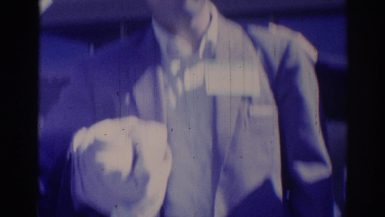 Two men in suits stand close indoors, one holding a cup or cloth near the camera.