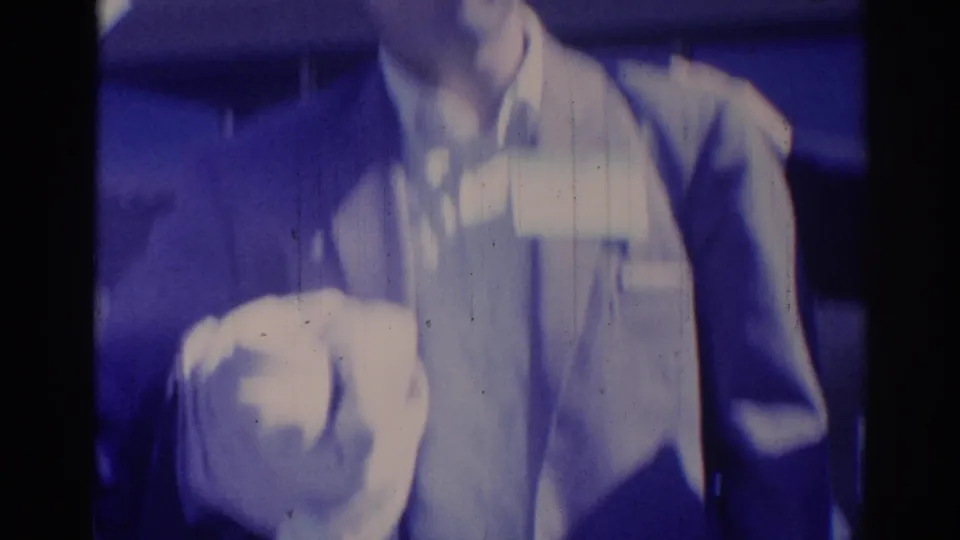 1967 Los Angeles home-movie frame of suited men standing close in a blue-cast indoor event setting.