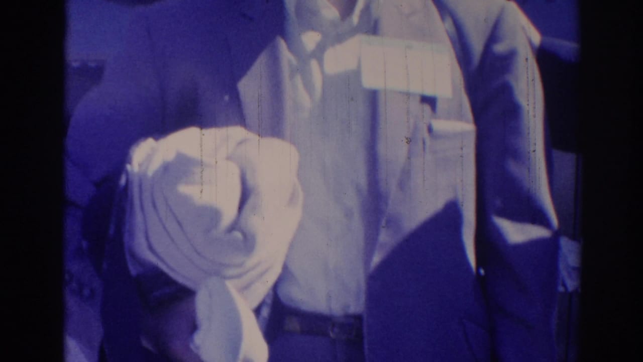 Blue-tinted 1967 Los Angeles frame of a man in a jacket with a name tag at a crowded indoor gathering.