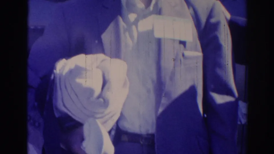 Blue-tinted 1967 Los Angeles frame of a man in a jacket with a name tag at a crowded indoor gathering.