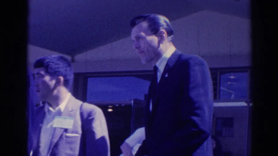 1967 Los Angeles frame showing a younger man with a name tag near an older suited man during conversation.