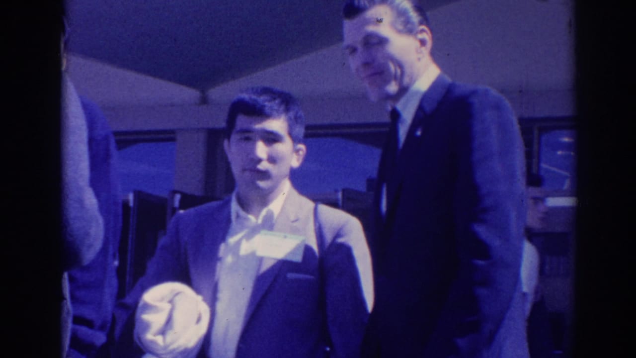 1967 Los Angeles 8mm frame of a younger man holding a light garment while an older man in a dark suit stands nearby.