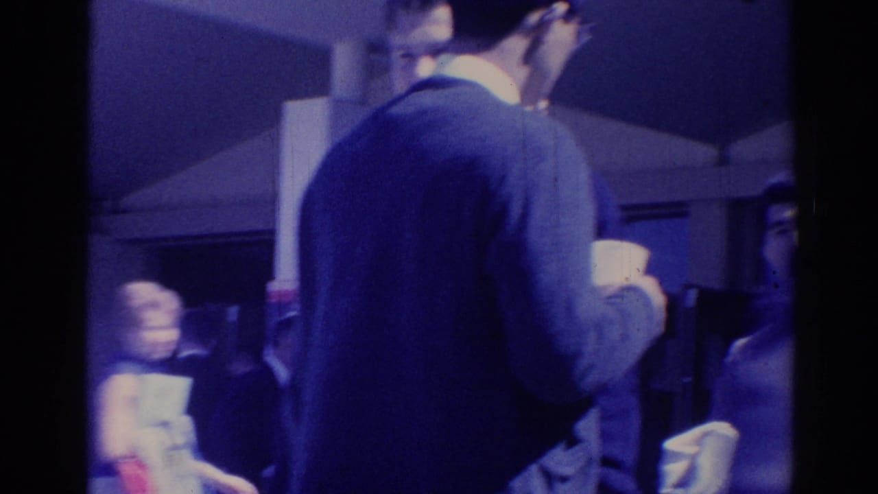 Candid 1967 Los Angeles frame of a man in a sweater drinking from a cup while people pass in the background.