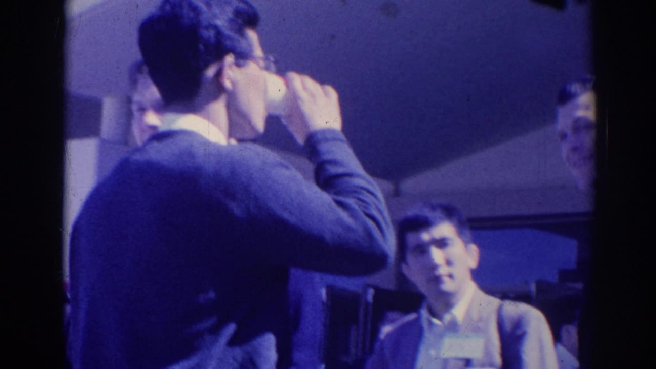1967 Los Angeles home-movie frame with a man drinking from a cup as suited attendees look on during a gathering.