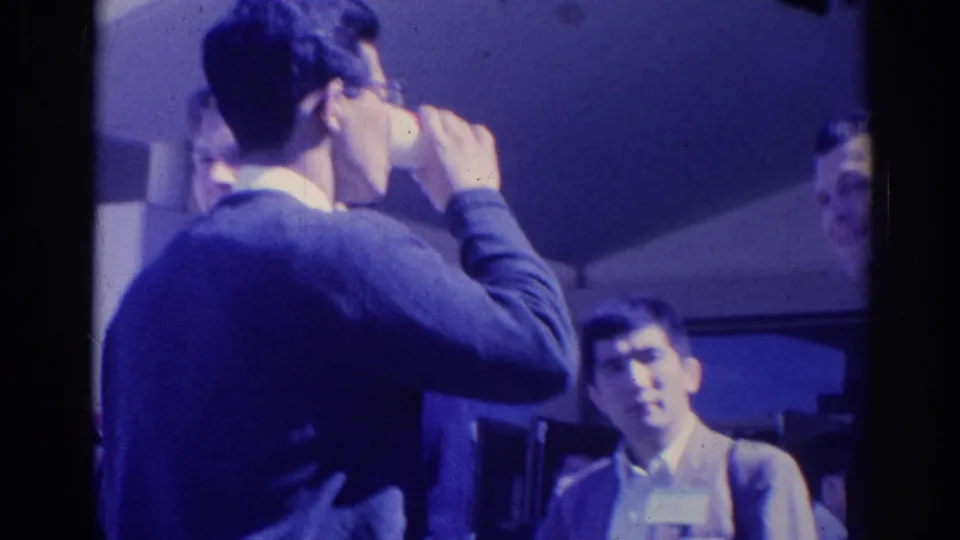 1967 Los Angeles home-movie frame with a man drinking from a cup as suited attendees look on during a gathering.