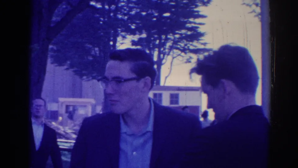 1967 Los Angeles 8mm frame showing two young men in dark jackets talking outdoors, with trees and distant people behind them.