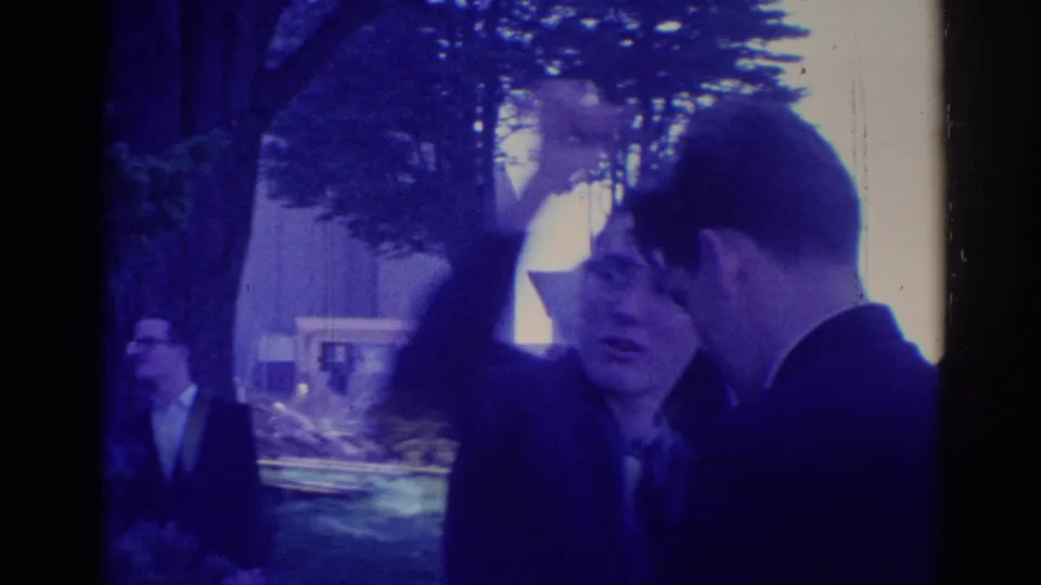1967 Los Angeles home-movie frame of a young man with glasses raising his arm while speaking with another man in a suit.