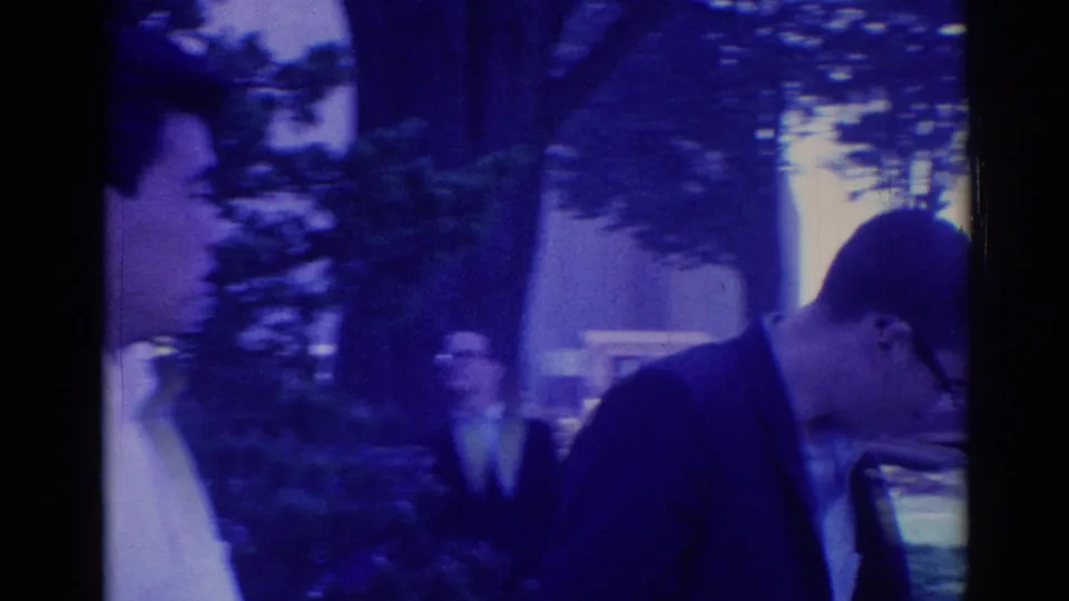 1967 Los Angeles home-movie frame showing suited young men walking outdoors near trees, a sidewalk, and a parked car.