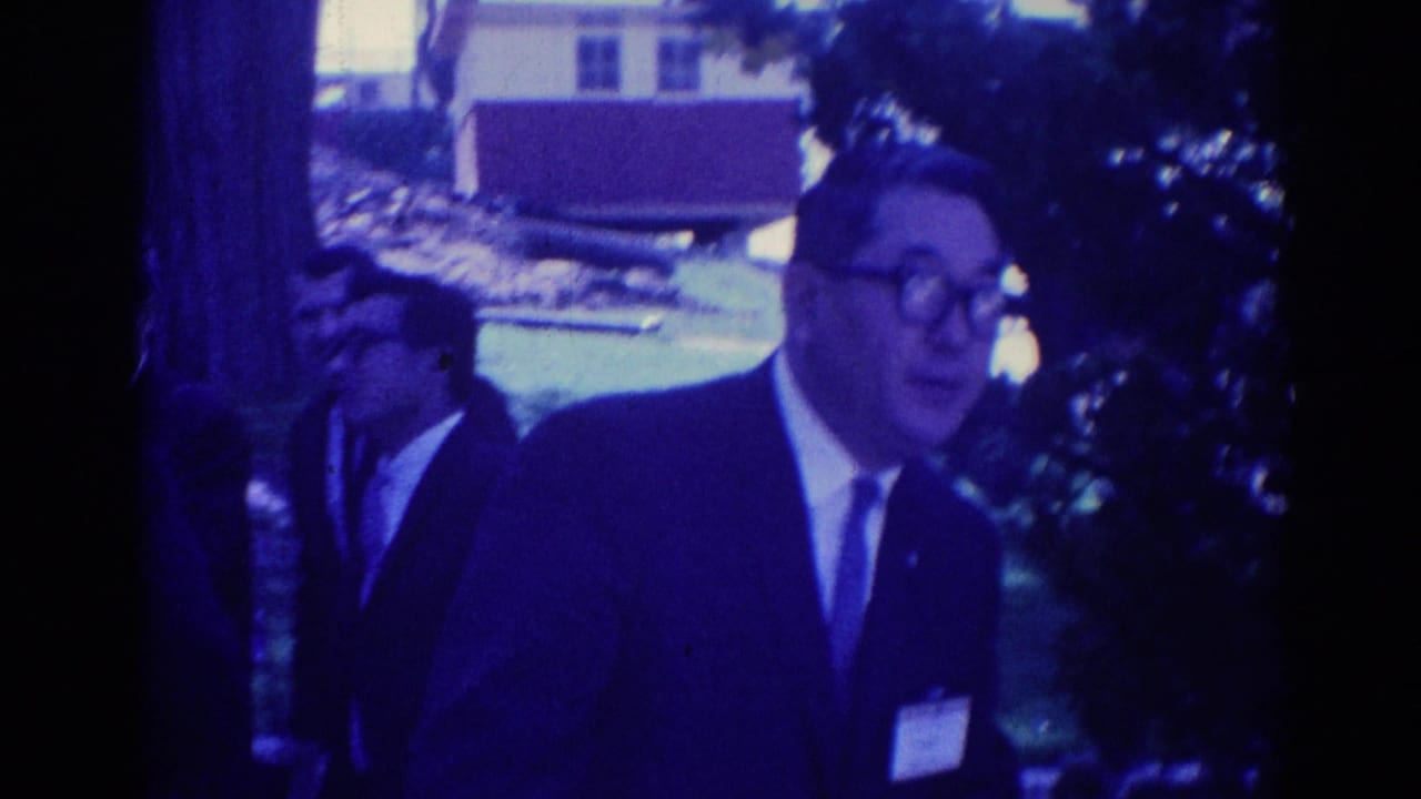 Suited men walk outdoors near trees and a building, with one man in glasses closest to camera.