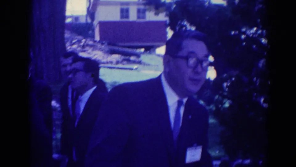 1967 Los Angeles home-movie frame showing men in dark suits walking through a landscaped outdoor area near a small building.