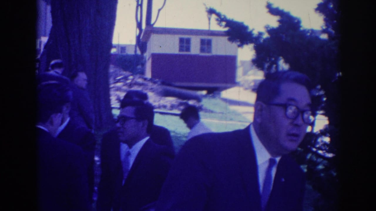 Men in suits appear to move together outdoors in Los Angeles, with trees, grass, and a small building behind them.