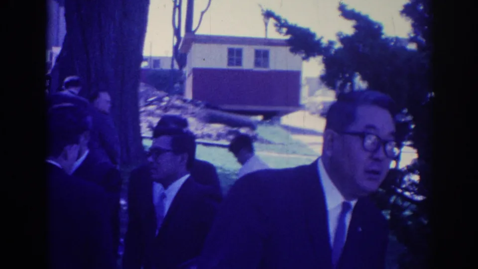 Men in suits appear to move together outdoors in Los Angeles, with trees, grass, and a small building behind them.