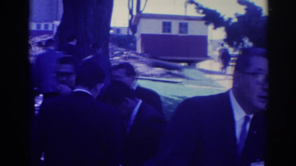 A handheld 8mm frame catches suited adults in a shaded landscaped area during an apparent formal outdoor gathering.