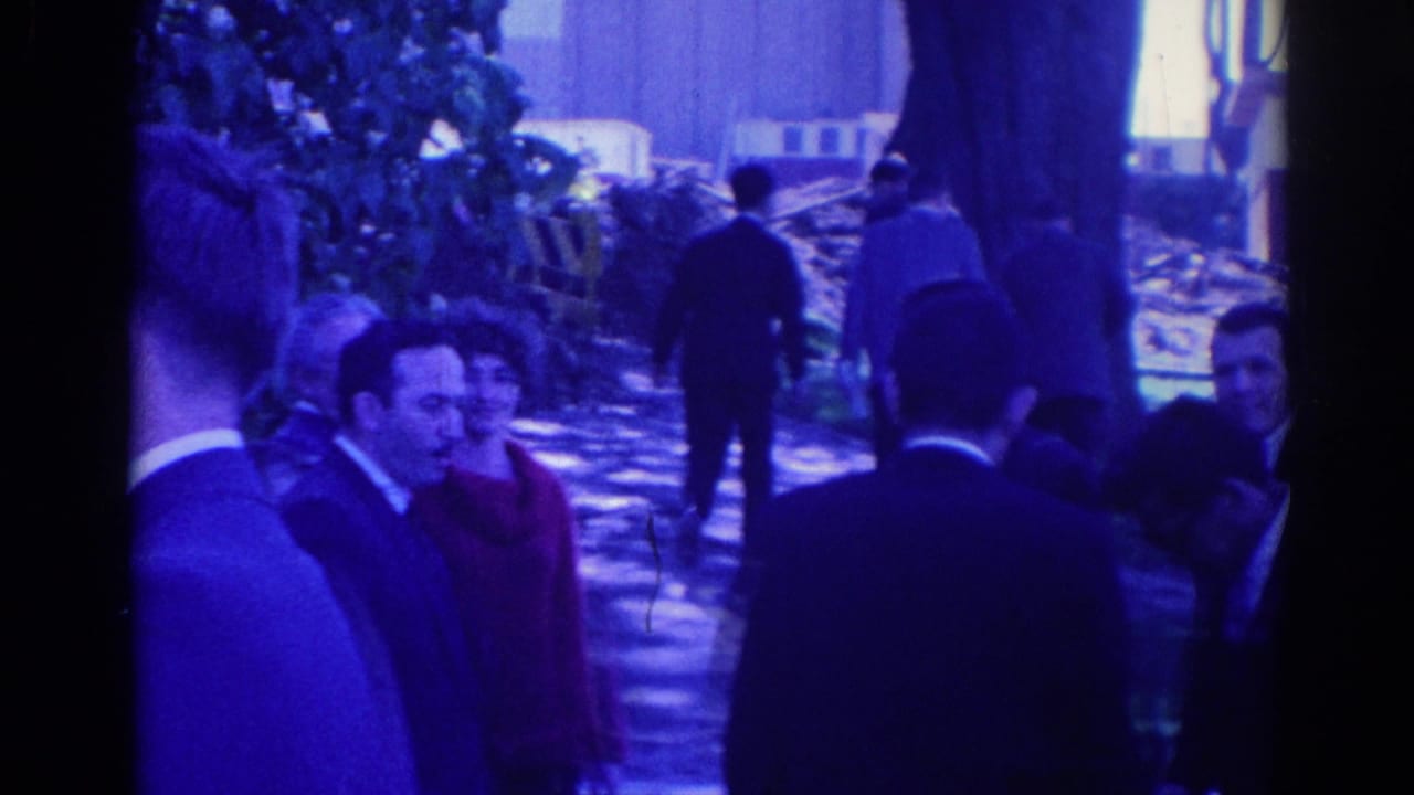 Adults in formal clothing gather outdoors among trees near a building.