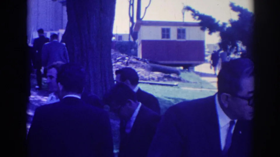 Blue-cast 8mm footage shows formally dressed adults near a tree and walkway during the Los Angeles wake gathering.