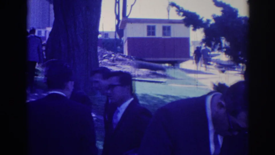A 1967 Los Angeles home-movie frame shows suited men in quiet conversation beside trees, with other adults walking in the background.