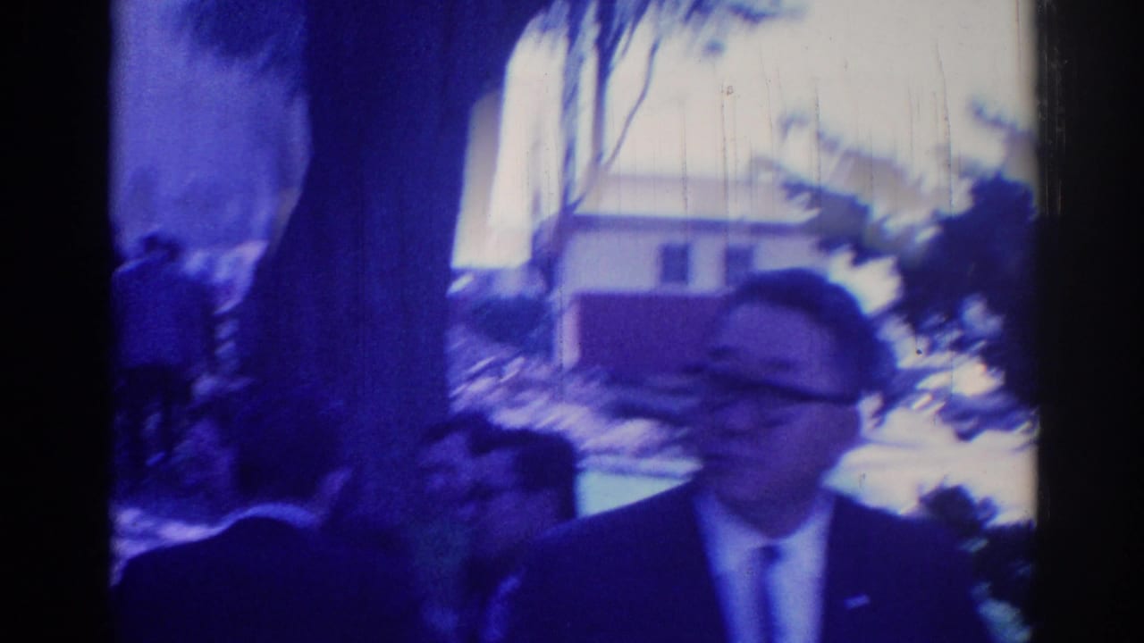 Suited men outside a building, with one man laughing in soft blue-tinted 8mm footage.