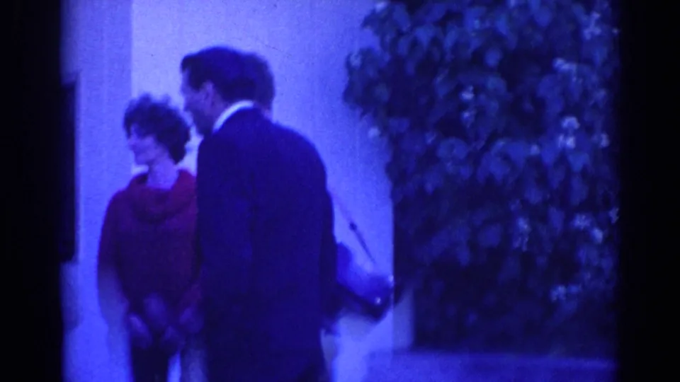 1967 Los Angeles frame of a woman in a red top and a suited man conversing near an exterior wall and shrubs.