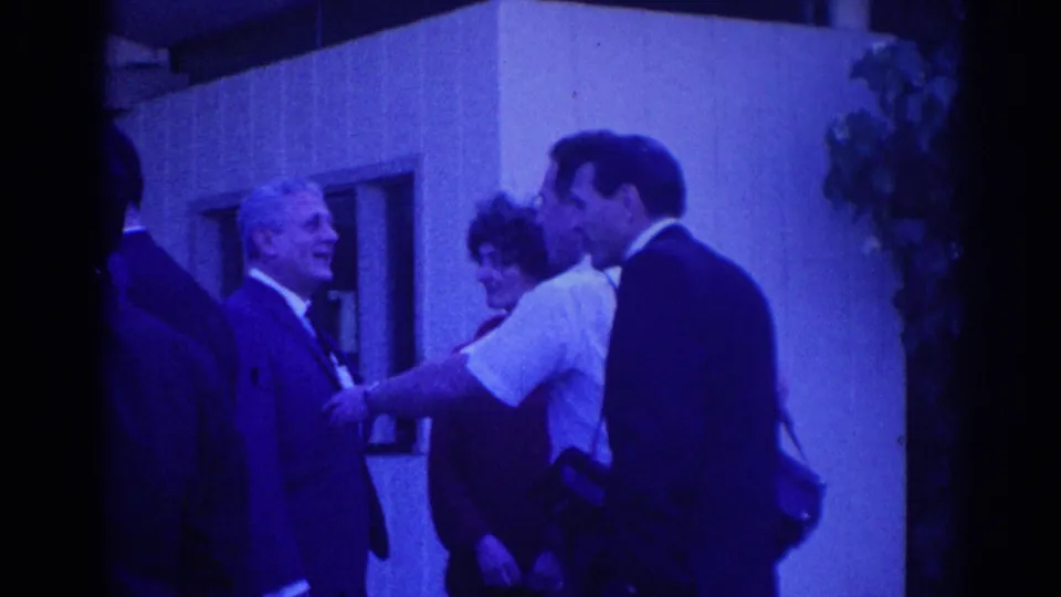 1967 Los Angeles home-movie frame with suited adults and a woman nearby during an outdoor greeting or handshake.