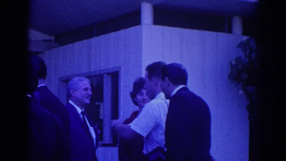 1967 Los Angeles home-movie frame of suited men and a woman gathered outside a modern building, with one man turned toward the group.