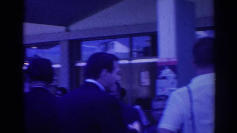 Men in suits and a man in a white short-sleeved shirt stand outside a Los Angeles building with glass windows and partial signage.