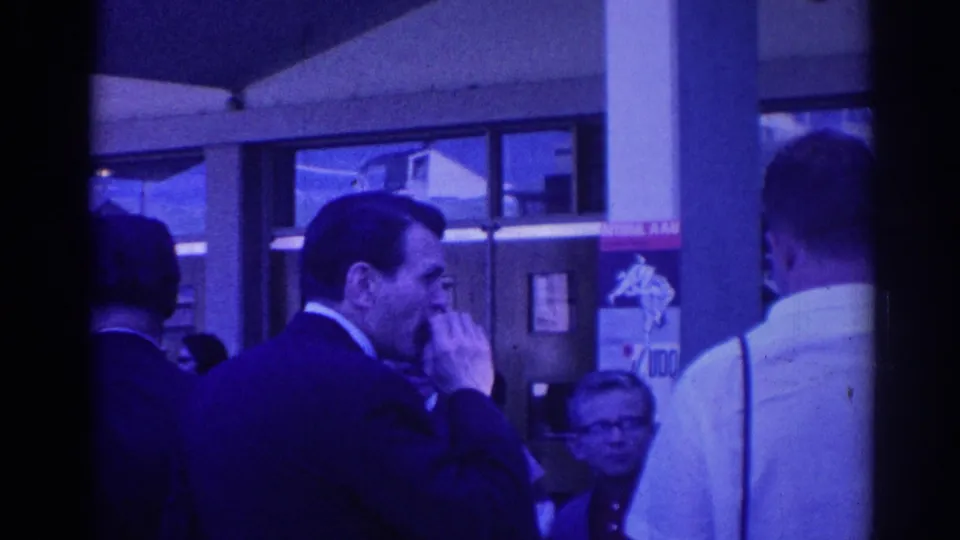1967 Los Angeles frame showing a suited man smoking a cigarette among adults outside a windowed building with a man in a white shirt nearby.