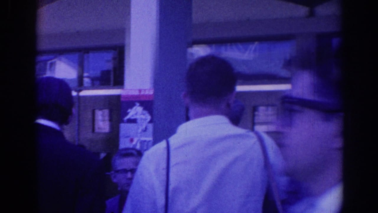 Adults stand outside a building, with a man in a white shirt in the foreground and posters on the wall behind them.